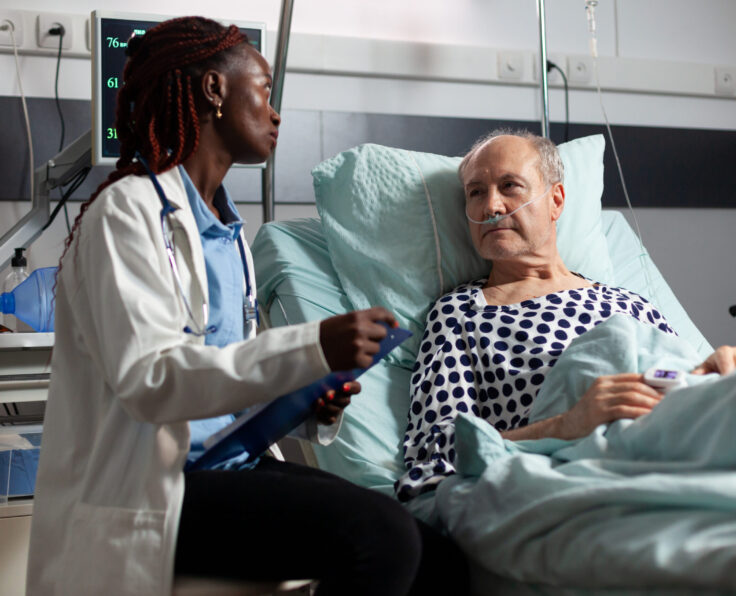 African american doctor reading diagnosis from clipboard to sick ill unwell senior patient laying in bed, breathing with help from oxygen mask, listening discussing with medical staff about recovery.